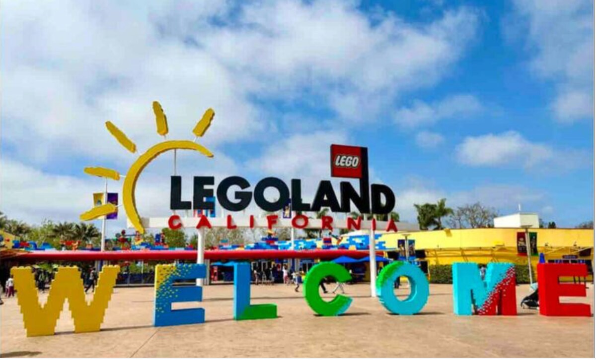 The entrance to Legoland California is marked by a large, colorful sign featuring the word 'WELCOME.' Bright letters in various colors create an inviting atmosphere, while a large sun emblem rises above the entrance. A clear sky with fluffy white clouds complements the festive scene.