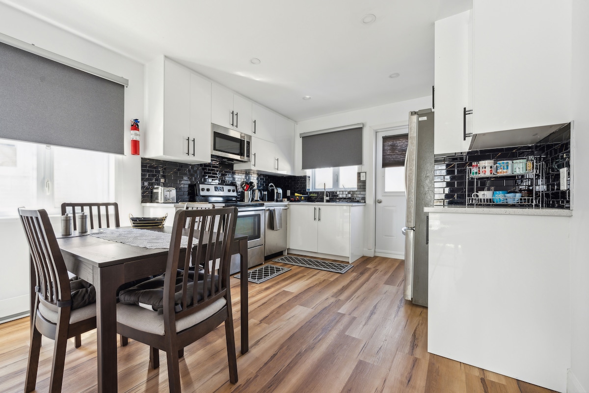 A modern kitchen is shown, featuring sleek white cabinetry and a dark tiled backsplash. Stainless steel appliances are visible, along with a dining table surrounded by four wooden chairs. Natural light enters through large windows with gray roller shades, illuminating the warm wooden flooring.