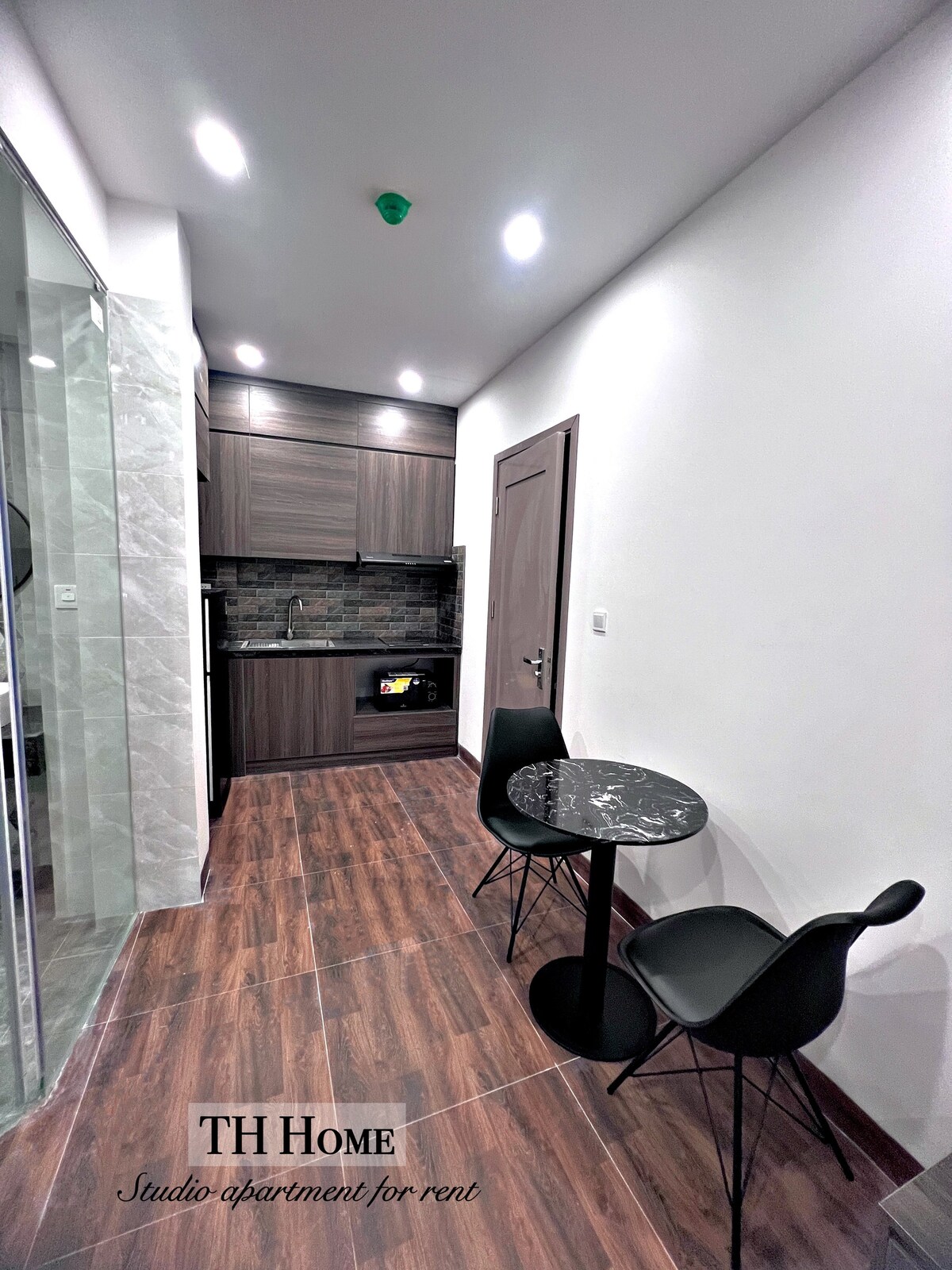 A studio apartment kitchen area is displayed, featuring sleek wooden cabinetry and modern appliances. A small marble-top table is set for two, complemented by black chairs. The warm wood flooring contrasts with the light walls, creating a cozy yet functional space.