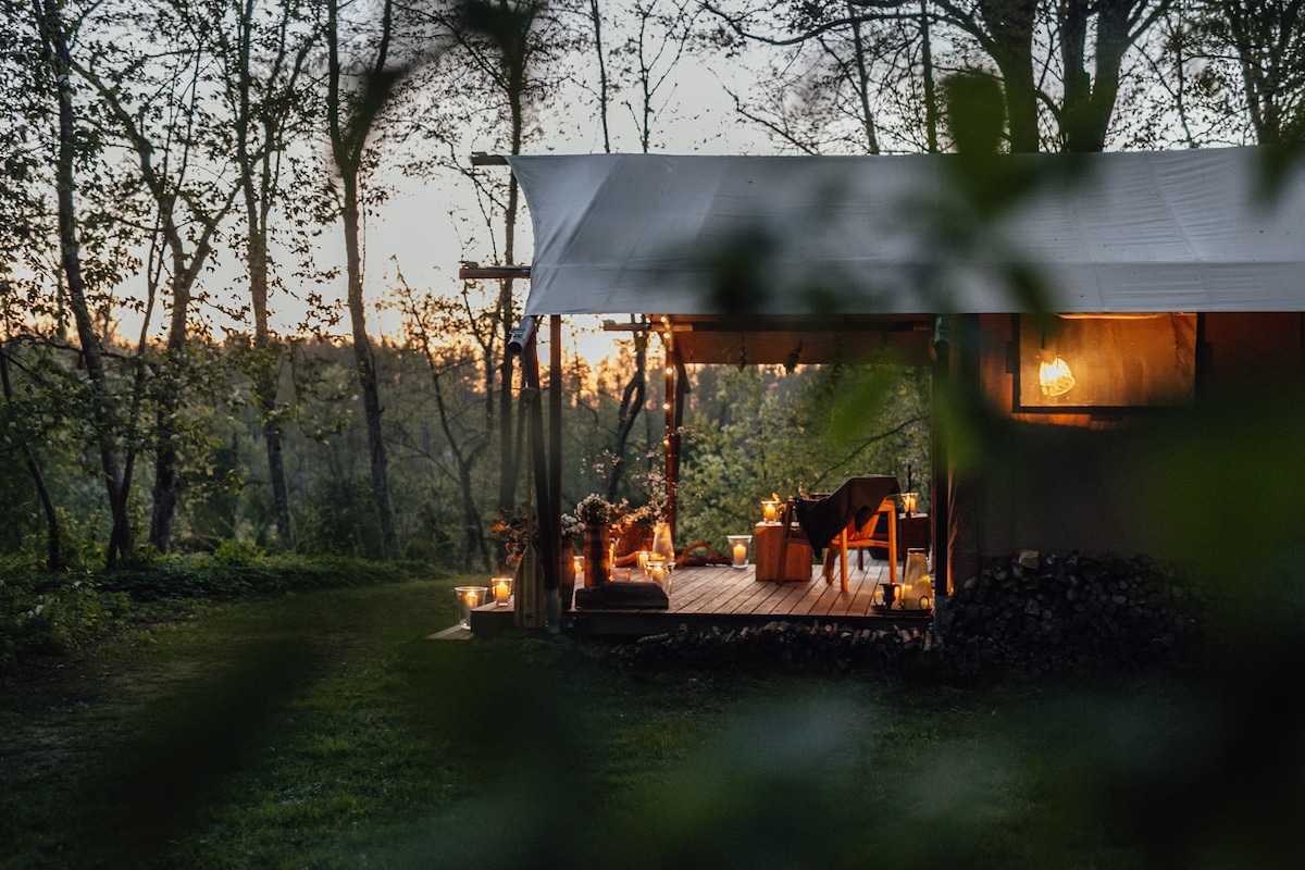 An outdoor glamping space is partially visible through foliage, with warm lighting illuminating a cozy wooden deck. The setting sun casts a soft glow over the surrounding trees, creating a serene atmosphere perfect for relaxation.