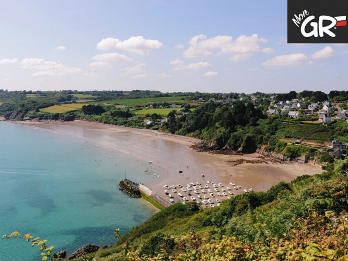 A scenic view of a sandy beach is framed by lush green hills under a bright blue sky dotted with clouds. White boats are anchored along the shore, while gentle waves touch the coastline, creating a serene atmosphere.