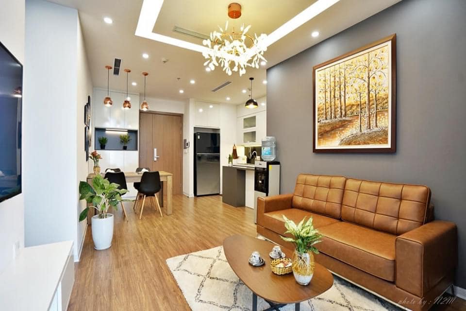 A modern living area features a brown leather sofa, a decorative coffee table, and a vibrant wall art piece. Natural light fills the space through a large window, and a small dining table is set nearby. Potted plants add a touch of greenery.