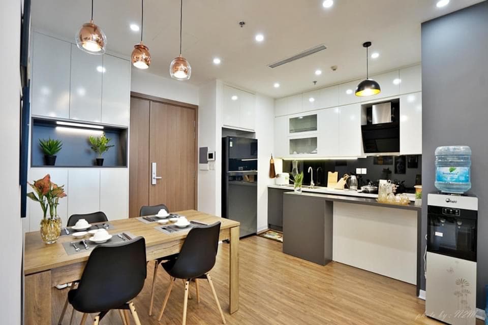 A modern kitchen and dining area is illustrated, featuring a wooden dining table with black chairs. The kitchen is equipped with sleek cabinetry, a stovetop, and a countertop. Soft lighting creates a welcoming ambiance, and decorative plants add a touch of greenery.