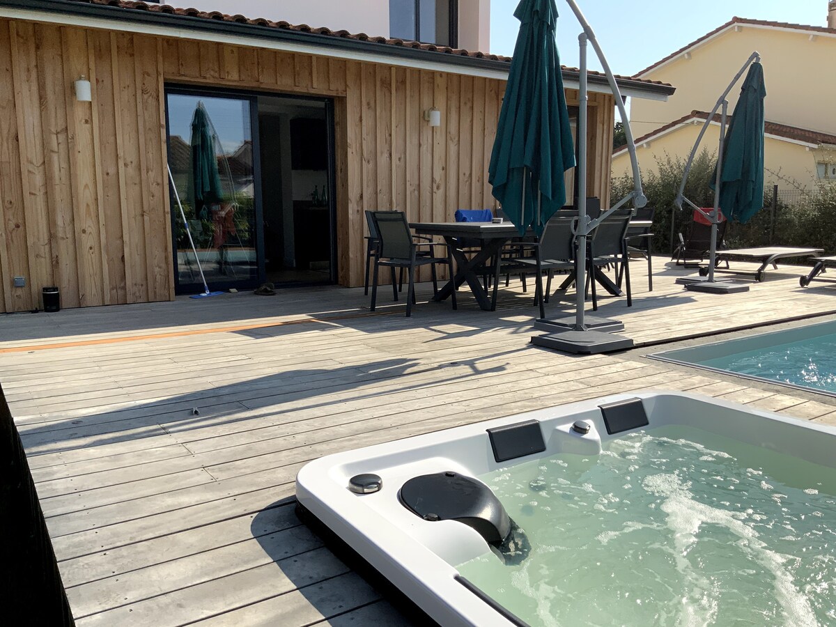 An outdoor terrace features a wooden deck surrounding a swimming pool and a jacuzzi. The jacuzzi accommodates six people and is positioned near seating arrangements. Umbrellas provide shade while the adjoining entrance leads into the house, enhancing outdoor relaxation.