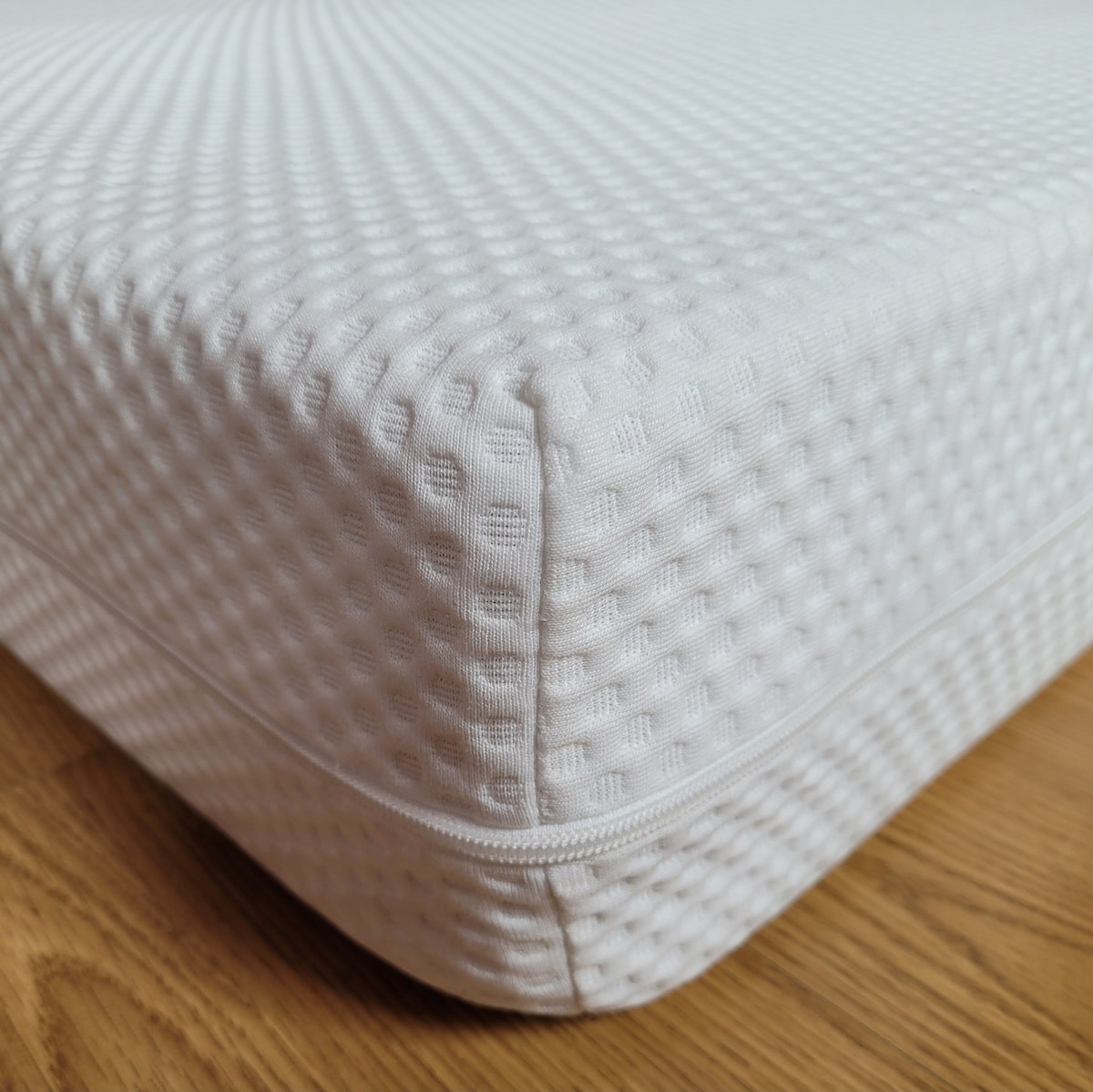 The image displays a close-up view of a white mattress with a textured surface. The corner of the mattress is clearly shown, emphasizing its cushioned design and zipper detail along the edge. The background features a warm wooden floor.