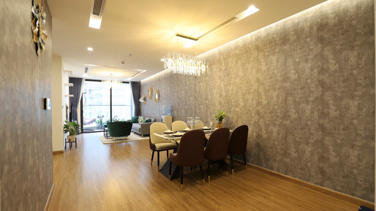A spacious dining area is featured with a round table surrounded by four elegant chairs. A modern chandelier illuminates the scene, complementing the subtle textured wallpaper. Natural light floods in from large windows, enhancing the inviting atmosphere of the open layout.