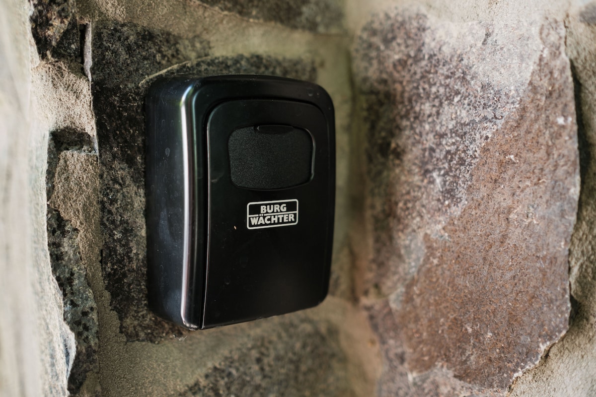 A black key box is mounted on a stone wall, featuring a secure clasp. The contrast between the smooth surface of the box and the rough texture of the surrounding stones is visible, highlighting the functional design intended for easy access to keys.