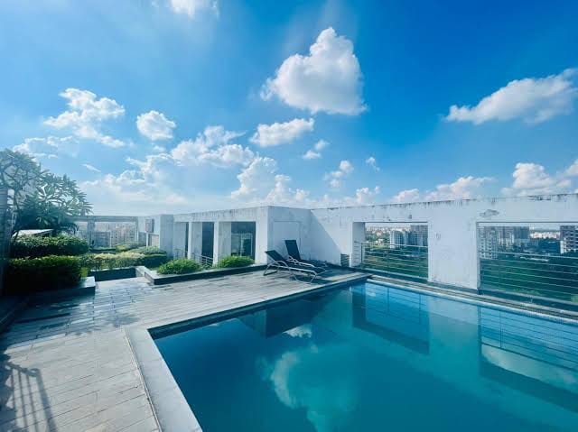 A rooftop pool area features a sleek, rectangular pool surrounded by a stone deck. Sun loungers are positioned nearby, offering a space for relaxation. The sky is bright blue with a few fluffy clouds, and lush greenery is present along the pool's edge.