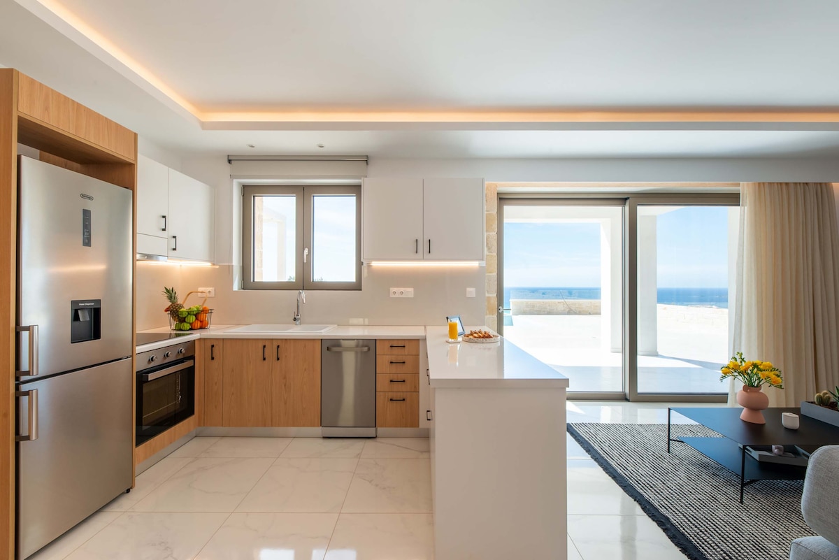 A modern kitchen is showcased with sleek cabinetry and high-end appliances, including a refrigerator and dishwasher. Natural light floods the space through large windows that offer views of the sea, complemented by a minimalist dining area and fresh fruit on the counter.