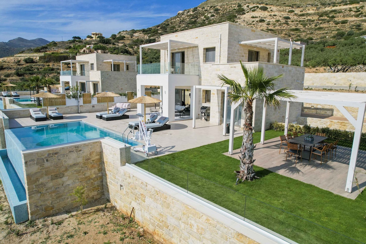 A contemporary villa complex is situated on a hillside, featuring two distinct structures with a spacious outdoor area. The area includes a private pool, sun loungers, and shaded dining spaces, surrounded by well-manicured gardens and stunning views of the surrounding landscape.