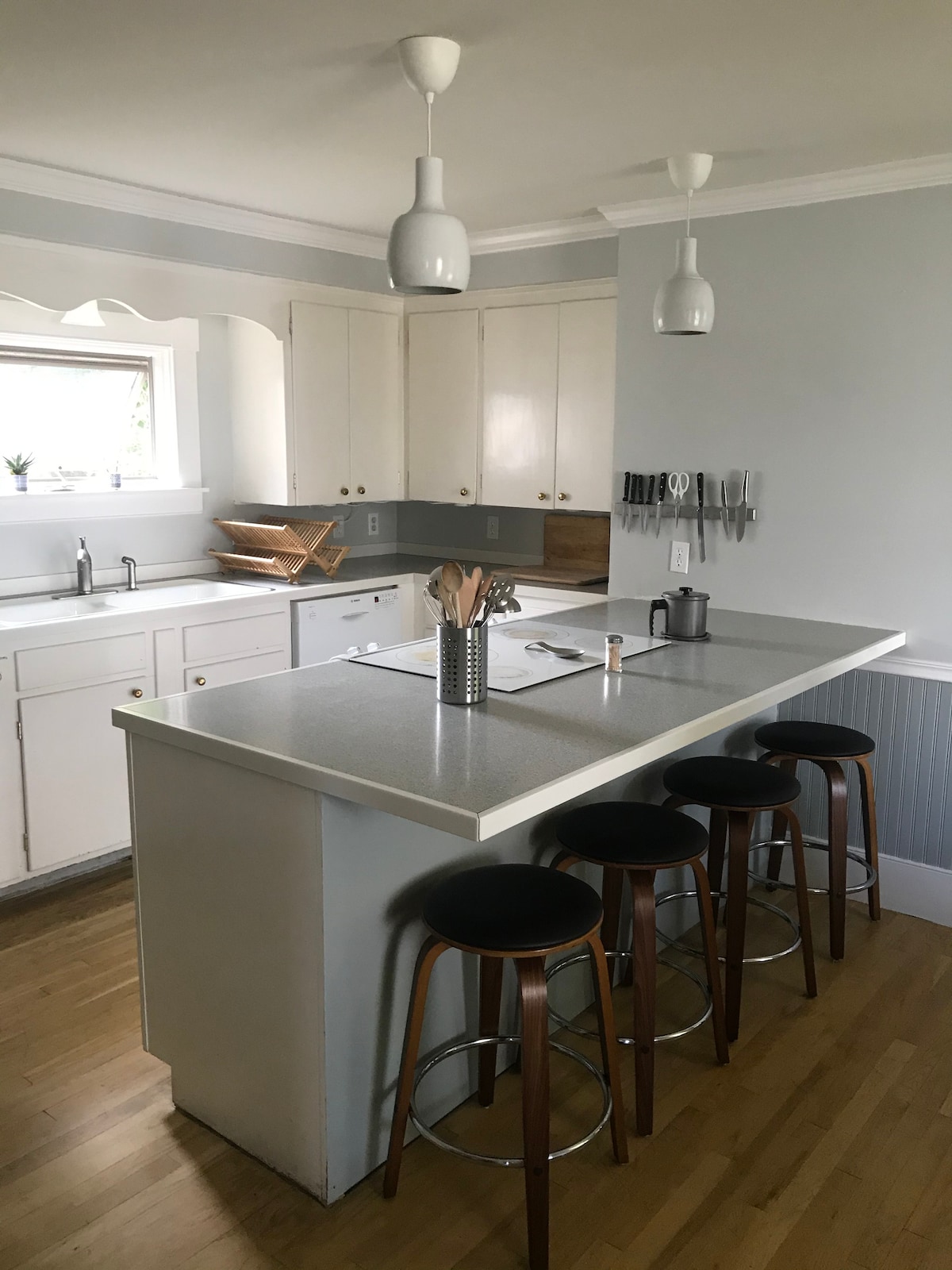 A spacious kitchen features modern fixtures and white cabinetry. A central island with seating for four stands at the forefront, surrounded by light wood flooring. Natural light enters through a window above the sink, complementing the sleek countertop and organized kitchen utensils mounted on the wall.