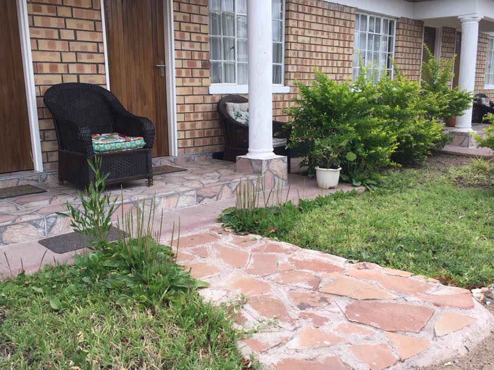 The entryway showcases a stone pathway leading to a covered porch. Two chairs are positioned to either side of the entrance, adorned with colorful cushions. Lush greenery surrounds the area, including potted plants and a small garden, providing a welcoming approach to the hotel.