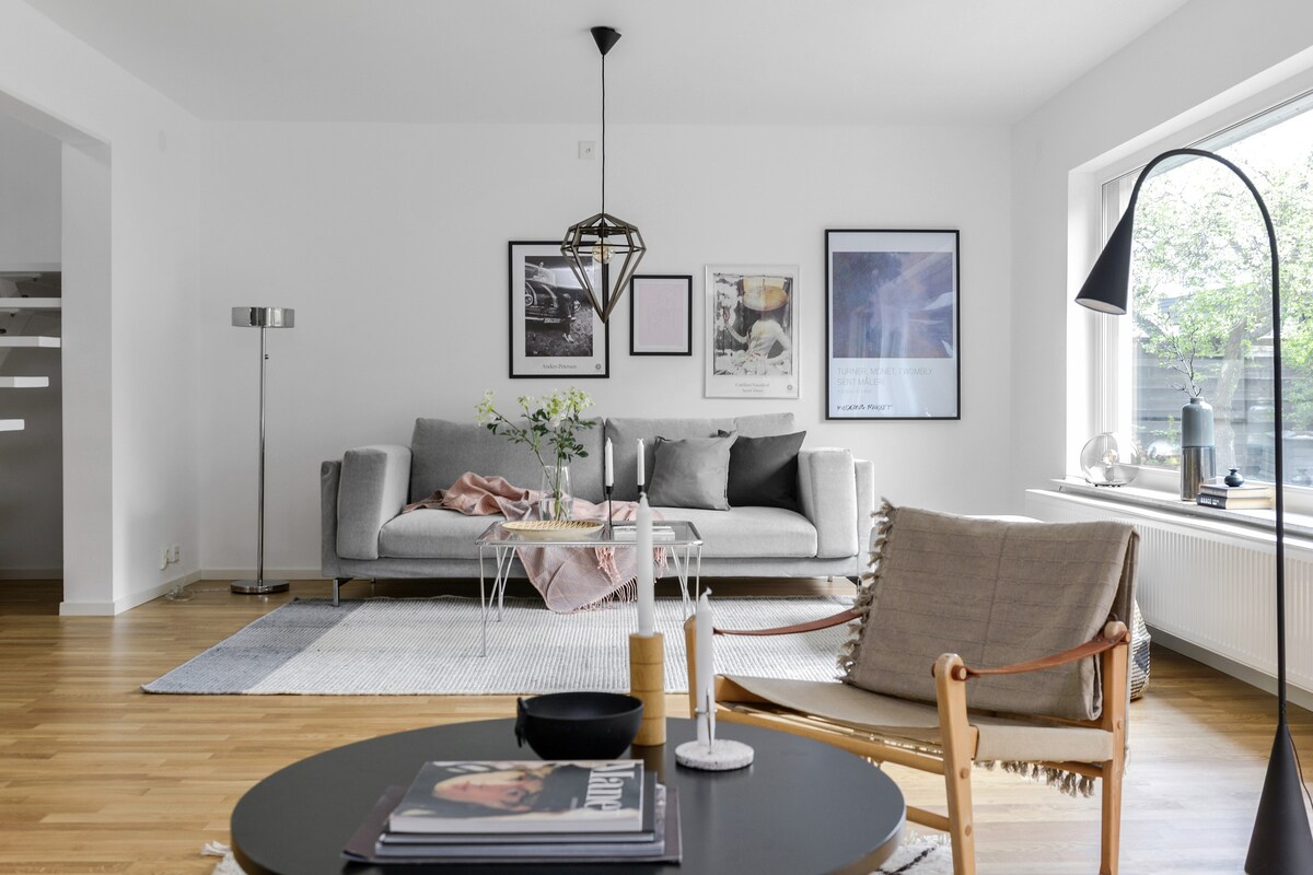 The living room features a grey sofa accompanied by a wooden armchair, each positioned around a coffee table. A floor lamp stands beside the sofa, enhancing the space, while natural light enters through a large window adorned with greenery outside.