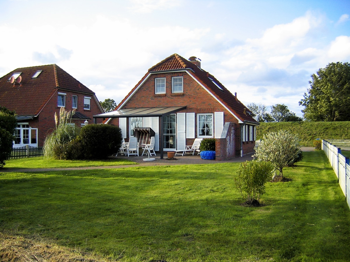 A two-story brick house is shown, featuring a welcoming facade with white shutters and a covered patio. Lush green lawns surround the property, with neatly trimmed hedges and shrubs. The nearby dyke serves as a scenic backdrop, enhancing the overall serenity of the setting.