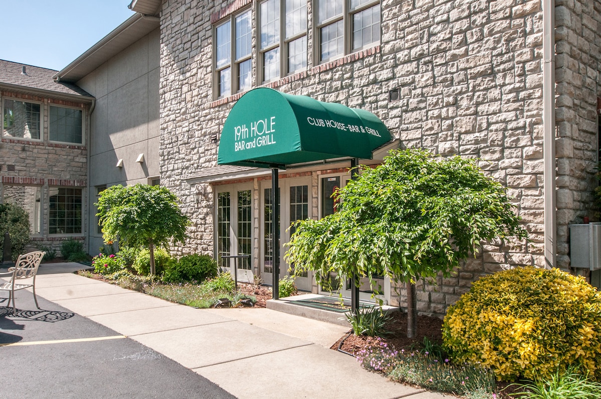 The entrance to the clubhouse features a green awning that reads '19th Hole Bar and Grill.' Flanking the entrance, neatly trimmed shrubs complement the stone-faced building, while a paved pathway leads to the doors. Outdoor seating is visible nearby.