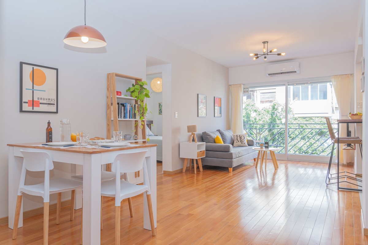 A well-lit living space features a comfortable gray sofa and a light wooden coffee table, surrounded by large windows that invite natural light. A dining table with casual white chairs, and a small workspace with a desk, enhance the functionality of the room.
