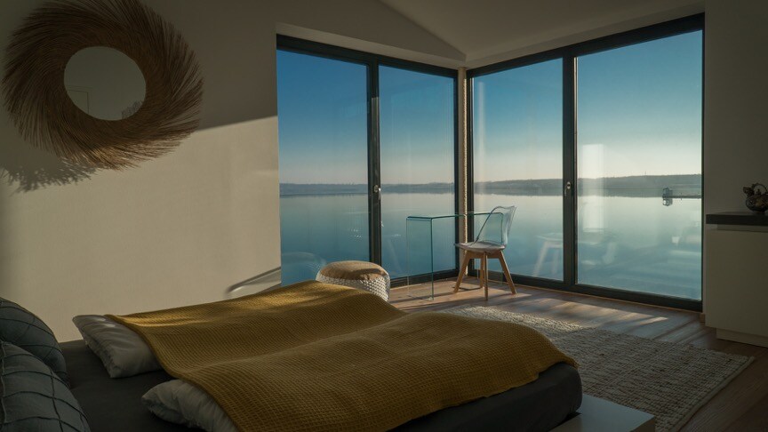 A spacious bedroom is presented, featuring a large bed with a mustard yellow blanket. Floor-to-ceiling windows offer panoramic views of the serene lake, allowing natural light to illuminate the room. A small glass table and a wooden chair are positioned near the windows.