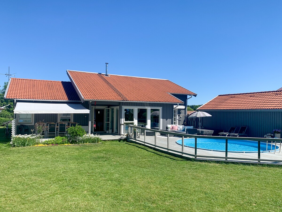 A single-story villa is shown with a red-tiled roof and gray exterior. Surrounding the property is a green lawn leading to a circular pool. Sun loungers are positioned nearby, and a spacious deck features outdoor seating, promoting relaxation and enjoyment of the sunny surroundings.