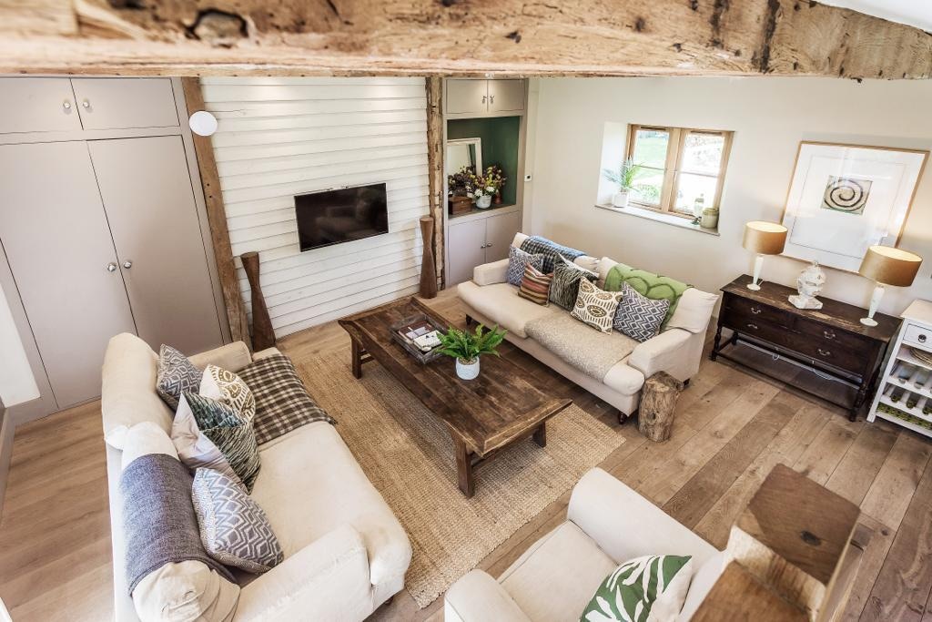 A spacious sitting room displays a blend of natural wood and soft textiles. Two comfortable sofas are arranged around a central wooden coffee table. A flat-screen TV is mounted on the wall opposite a sunlit window, while decor elements such as plants and artwork enhance the cozy atmosphere.