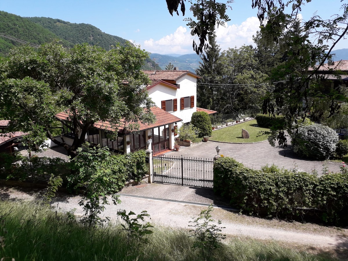 A charming accommodation is nestled amidst the lush greenery of the hills, featuring a two-story white building with red-tiled roofs. An inviting entrance gate leads to a gravel driveway, surrounded by trees and a well-kept garden that adds to the tranquil setting.