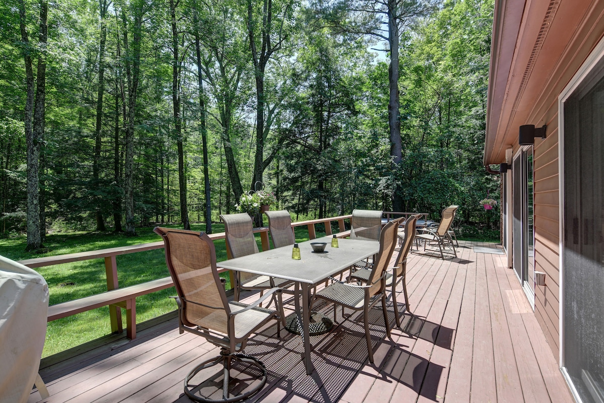 A spacious outdoor deck is furnished with a large dining table and multiple chairs, nestled among a backdrop of lush greenery. The area is designed for relaxation and dining, offering a peaceful view of the surrounding woods.