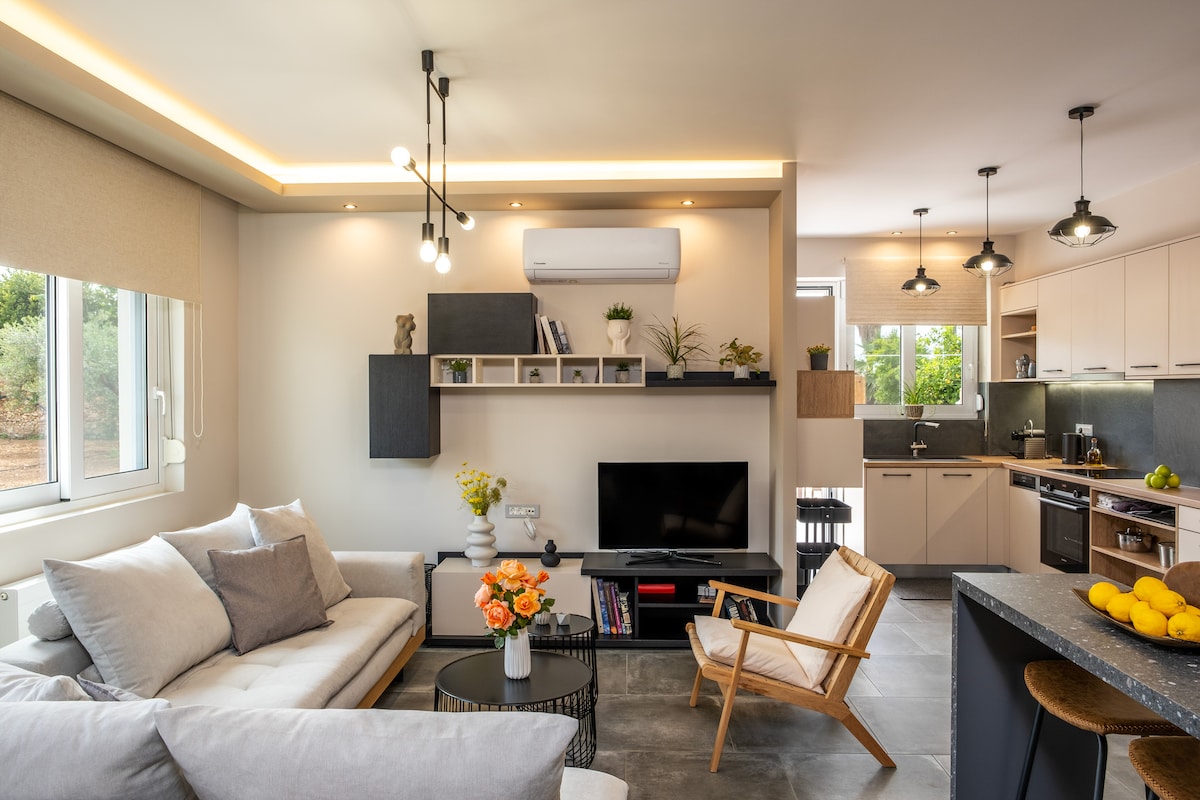 A modern living area features a light-colored sofa and wooden armchair, with a round coffee table displaying a bouquet of flowers. A television is mounted on the wall, and the kitchen is visible in the background with ample natural light from the windows.