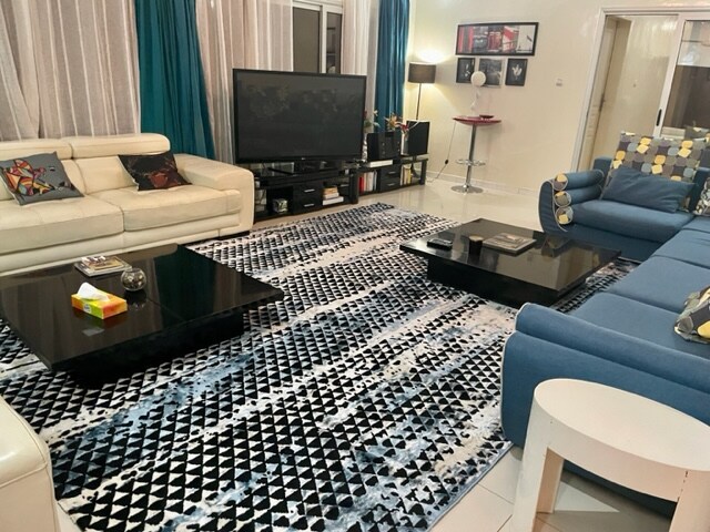 A spacious living area is highlighted by a patterned area rug that complements the modern furnishings. A large flat-screen TV is positioned on a low cabinet. Two coffee tables, a light-colored sectional couch, and a blue sofa provide ample seating, while sheer curtains allow natural light to filter through.