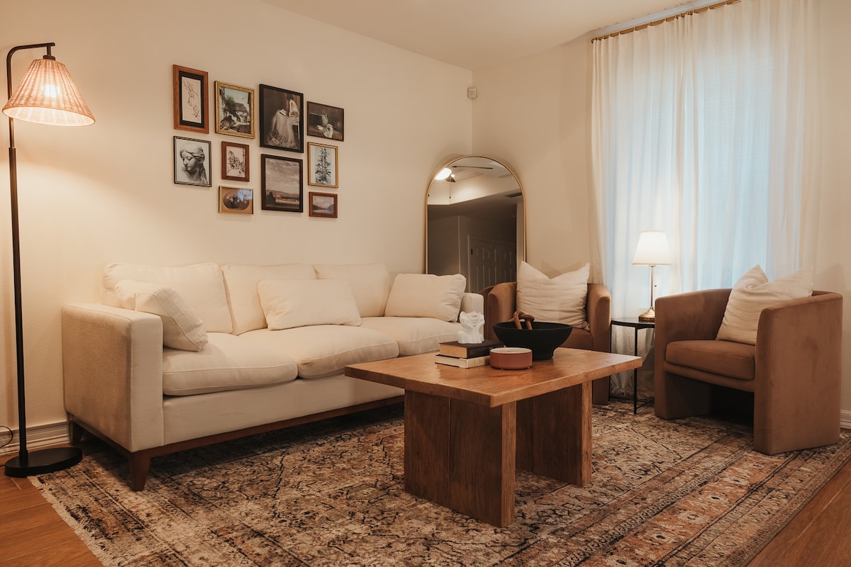 A comfortable living area features a light-colored sofa adorned with several cushions, complemented by two armchairs in soft beige. A wooden coffee table sits at the center, with a few books and a decorative bowl displayed on top. A gallery wall with framed images adds interest, while sheer curtains allow natural light to filter in.