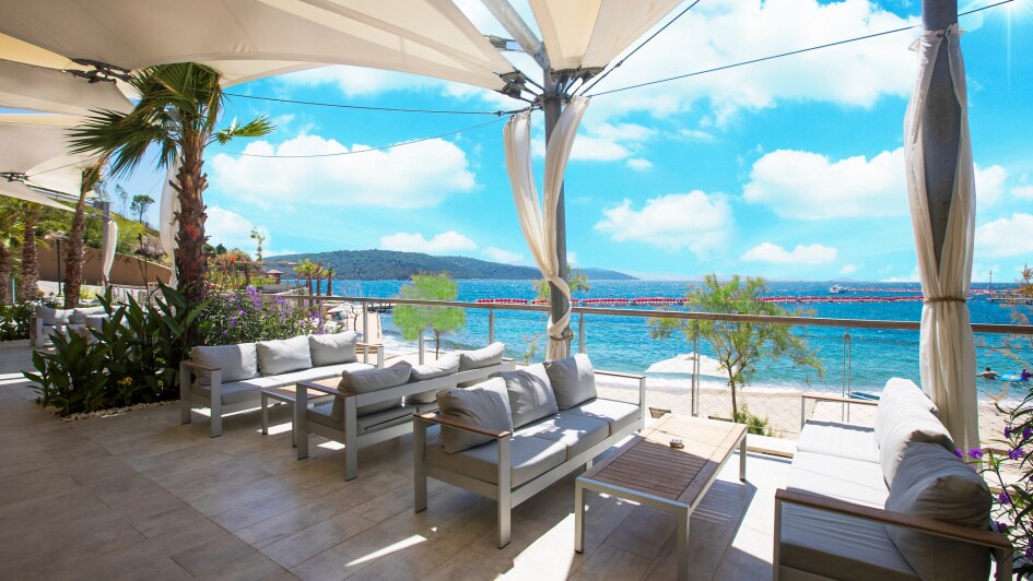 An outdoor lounge area features comfortable, cushioned seating overlooking the beach. Light fabric canopies provide shade, while palm trees and blooming plants add greenery. The picturesque view includes turquoise waters and a blue sky, enhancing the relaxing ambiance.