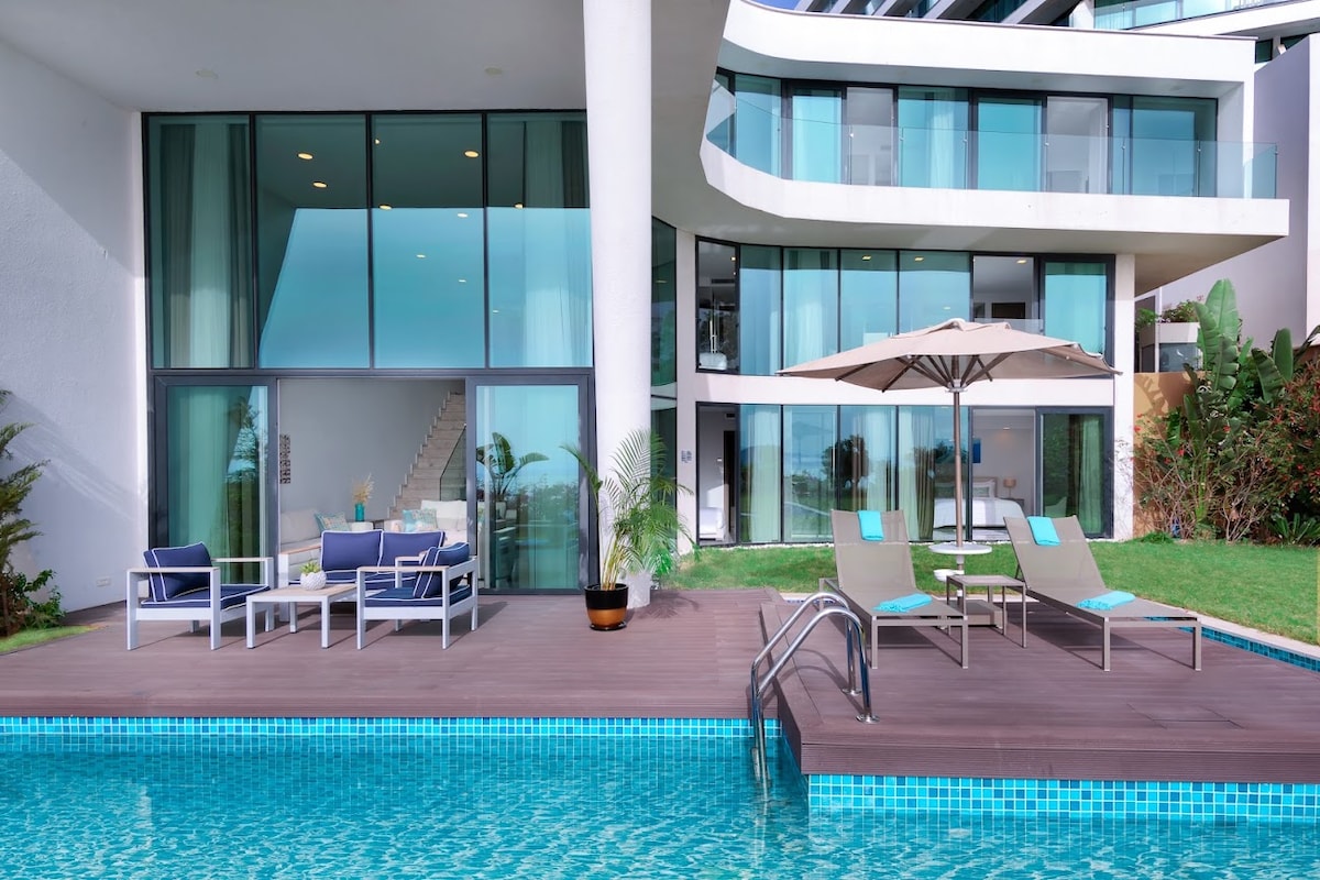 The outdoor area features a sparkling swimming pool bordered by a spacious wooden deck. Comfortable lounge chairs are arranged beside the pool, accompanied by a large umbrella for shade. Floor-to-ceiling glass doors lead into the modern building, enhancing the open and airy ambiance.