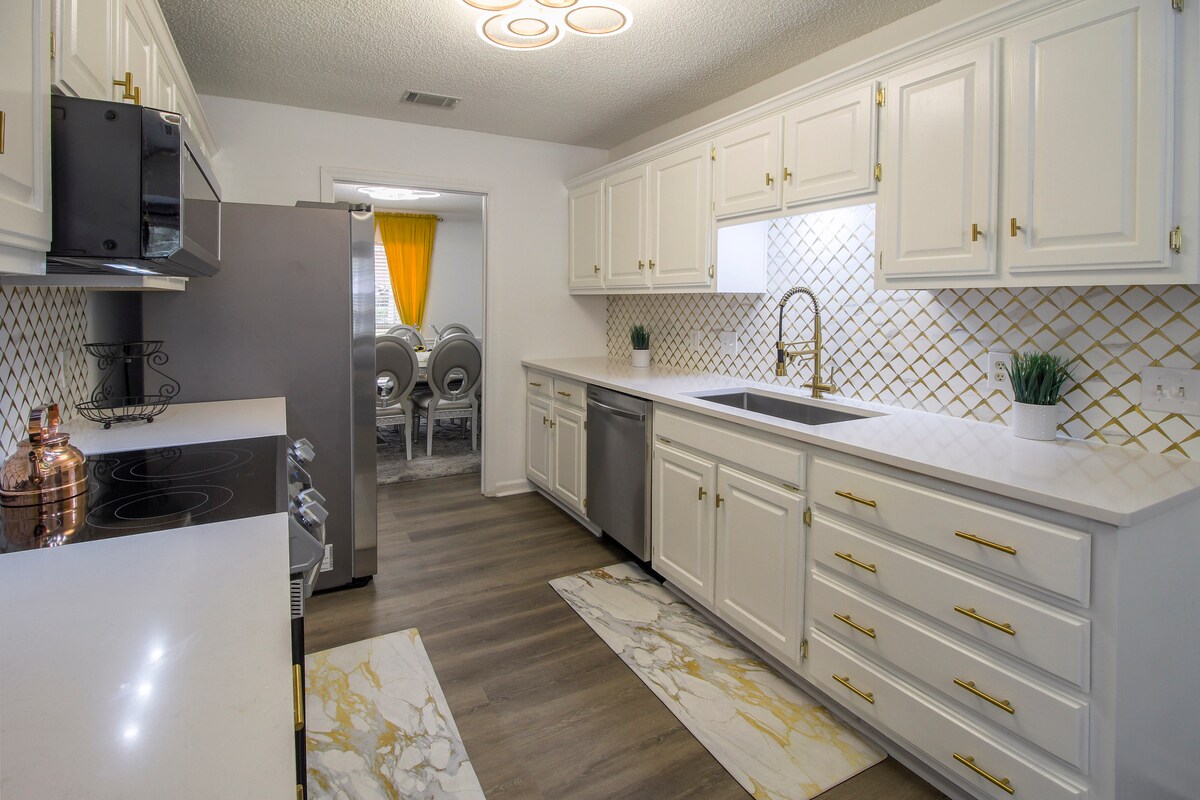 The kitchen features white cabinetry with golden hardware and a marble countertop, providing a clean and elegant appearance. Stainless steel appliances are included, and a modern sink is positioned under a window. Complementary area rugs add a touch of style to the space.