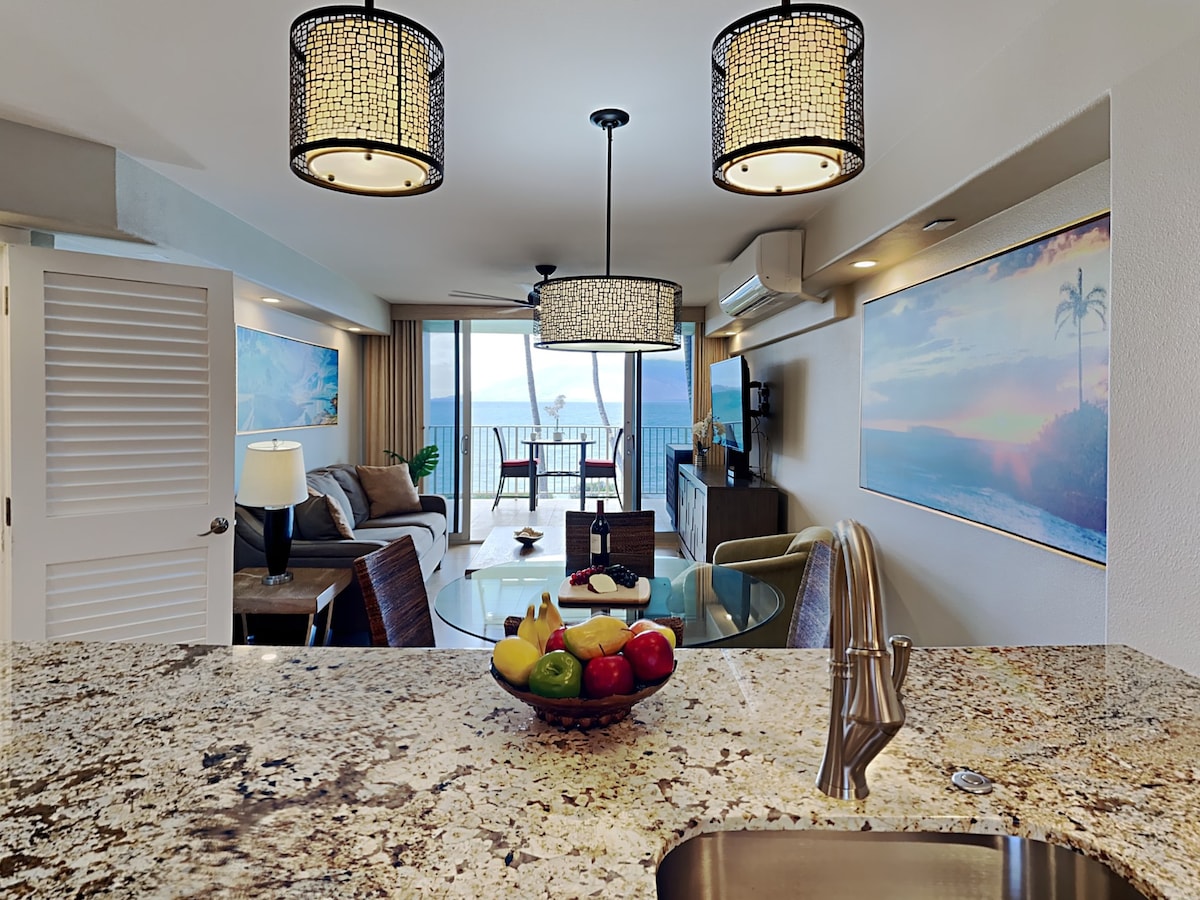 The kitchen area showcases a granite countertop, with a bowl of fresh fruit prominently displayed. In the background, a spacious living area features comfortable seating and a balcony with ocean views through large glass doors. Two elegant chandeliers provide ambient lighting.