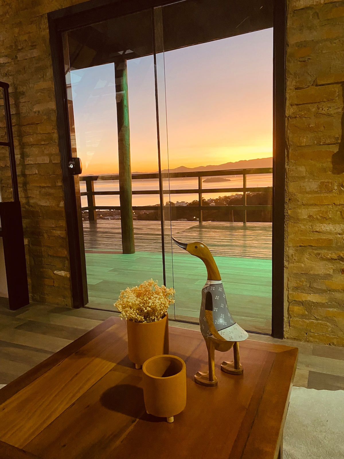 A wooden coffee table holds two decorative vases, one filled with flowers. In the background, a sliding glass door opens to a stunning sunset view over the lagoon, framed by a railing and distant mountains.