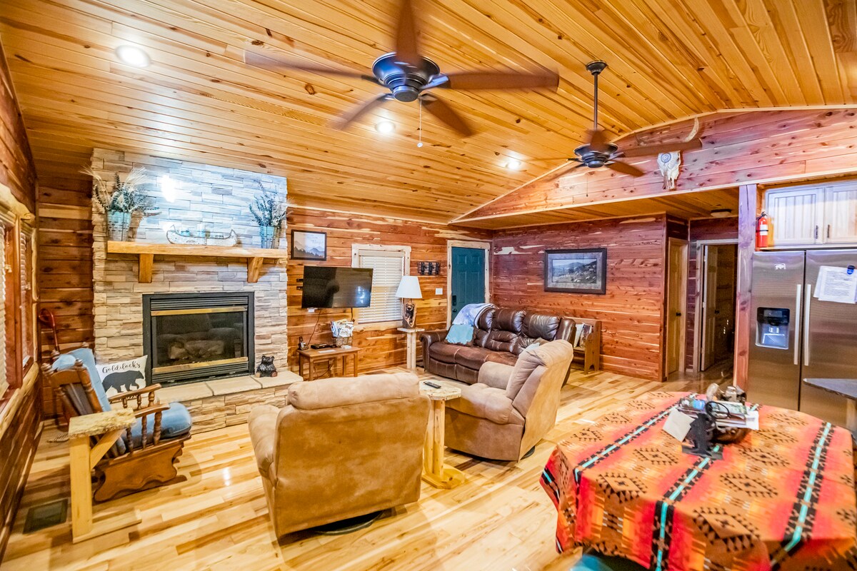 The living room features a stone fireplace, a comfortable sofa, and armchairs. A flat-screen TV is mounted above the mantel, and wood-paneled walls enhance the cozy ambiance. A vibrant patterned blanket is draped over a chair, while light illuminates the spacious area.