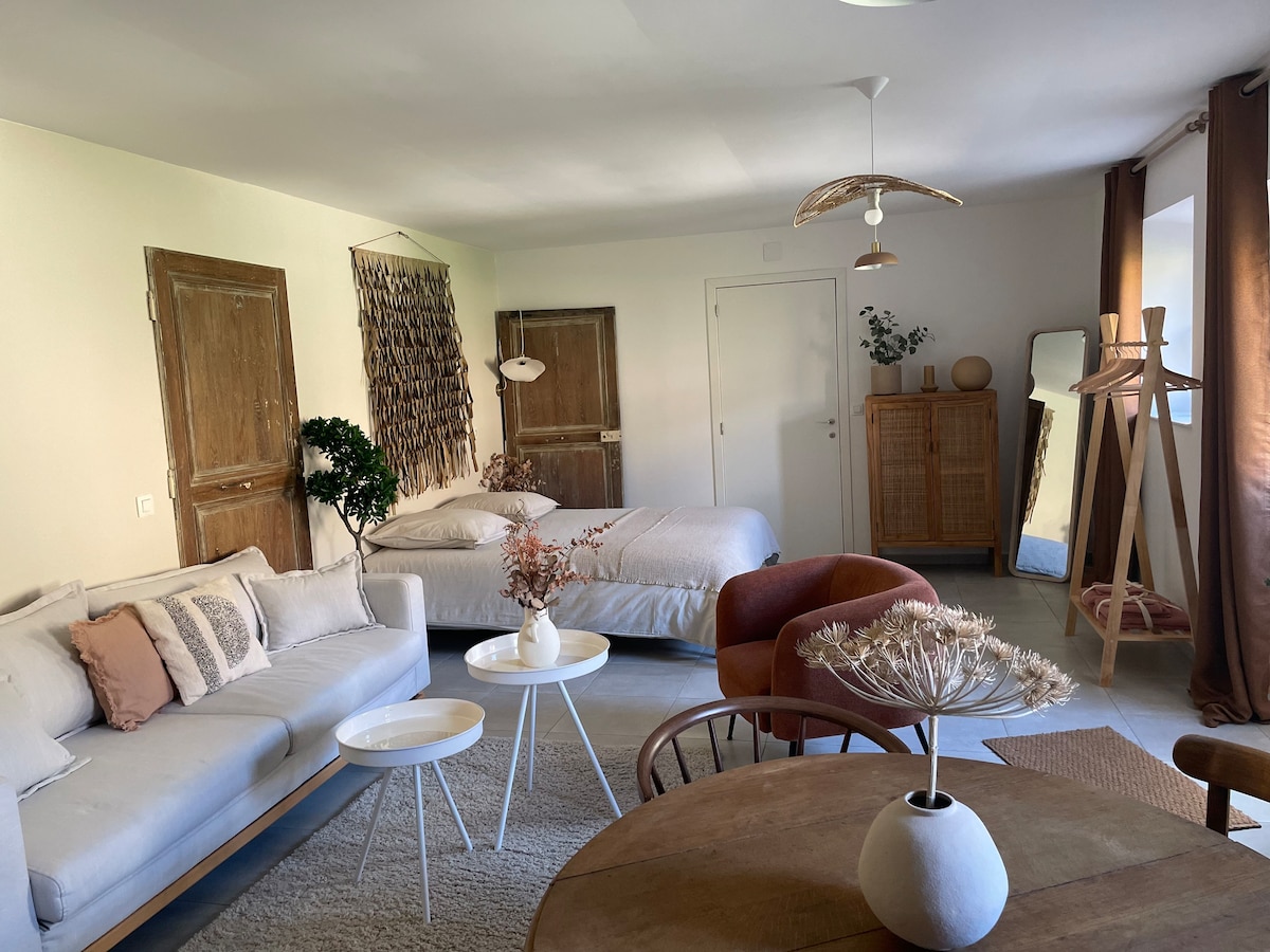 A light-filled studio features a spacious bed complemented by soft linens. A comfortable sofa and circular coffee table are present in the inviting living area. Natural elements are incorporated through decor, while wooden accents and plants enhance the warmth of the space.