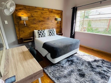 A cozy bedroom features a spacious bed with decorative pillows and a dark throw blanket. The wooden accent wall adds warmth, while natural light filters through the window, illuminating the room. A wooden desk is positioned alongside the bed, and a soft area rug lies beneath.