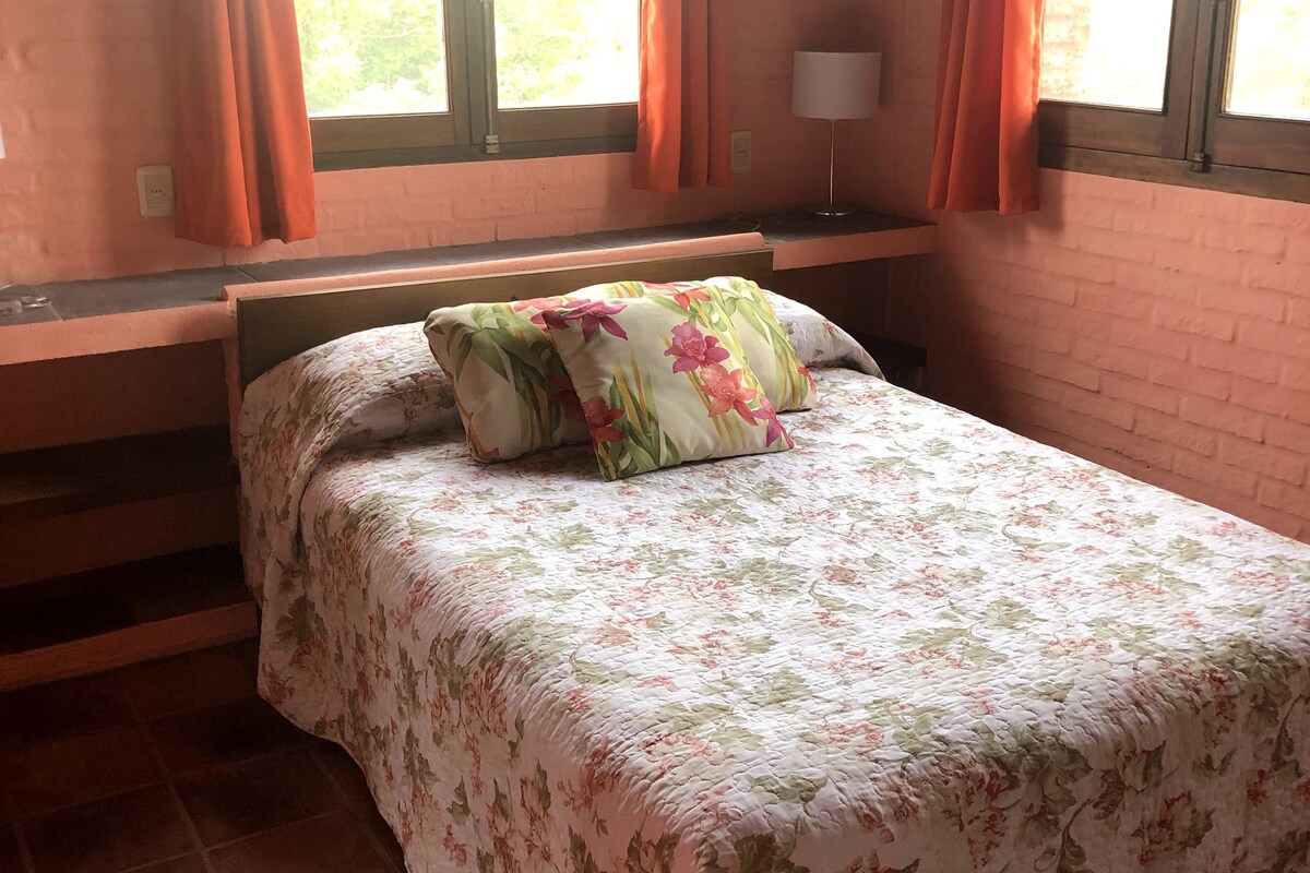 A comfortable sleeping area features a floral-patterned bedspread covering a spacious bed. A decorative pillow adds a touch of color. Natural light comes through large windows adorned with bright orange curtains, contrasting with the brick wall and creating a cozy atmosphere.
