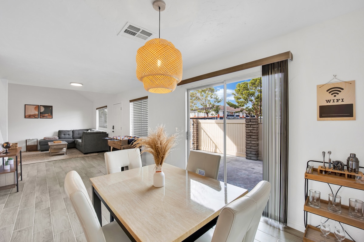 A well-lit dining area showcases a wooden table surrounded by four white chairs. A pendant lamp hangs above, providing soft illumination. In the background, sliding glass doors reveal a private patio, while a comfortable seating arrangement is visible in the adjacent living space.