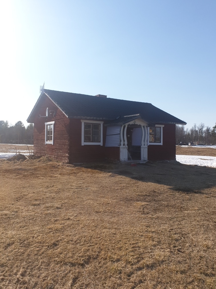 A charming red wooden house is set against a clear blue sky. The structure features a white door with decorative accents and several windows, allowing natural light to illuminate the interior. Snow patches are visible around the house, hinting at the changing seasons.