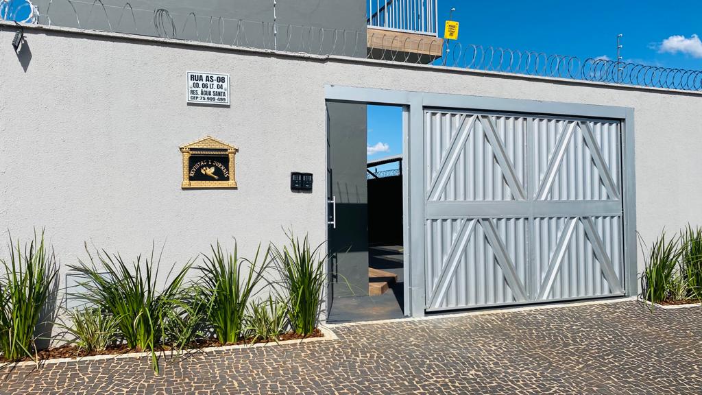 A secure entrance to the property is displayed, featuring a large, silver gate framed by low-growing green plants. A decorative plaque with a symbol is mounted beside the gate, which is set against a bright blue sky.