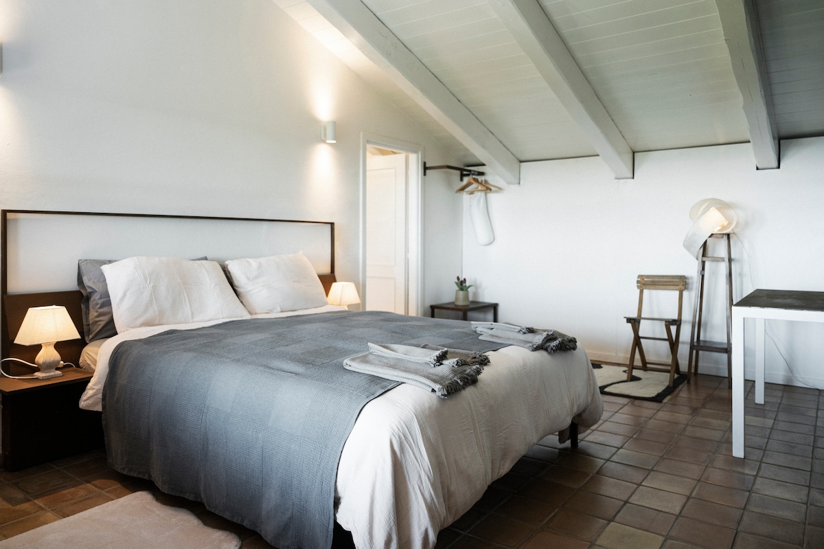 A well-lit bedroom features a spacious bed dressed in neutral bedding. Two bedside lamps provide soft illumination. A small table and a chair are positioned nearby, with an open closet visible in the corner. Exposed wooden beams add character to the ceiling.