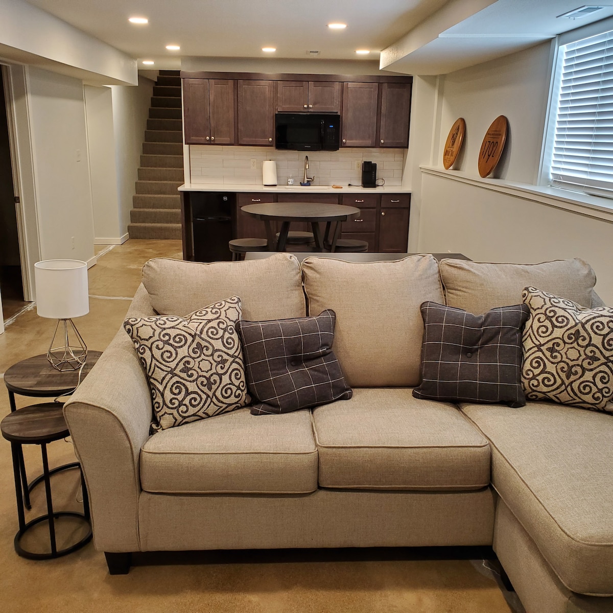 A spacious living area features a light-colored sectional sofa adorned with patterned throw pillows. A round dining table sits nearby, complemented by wood cabinetry that houses kitchen appliances. A staircase leads to the upper level, while soft lighting enhances the welcoming atmosphere.