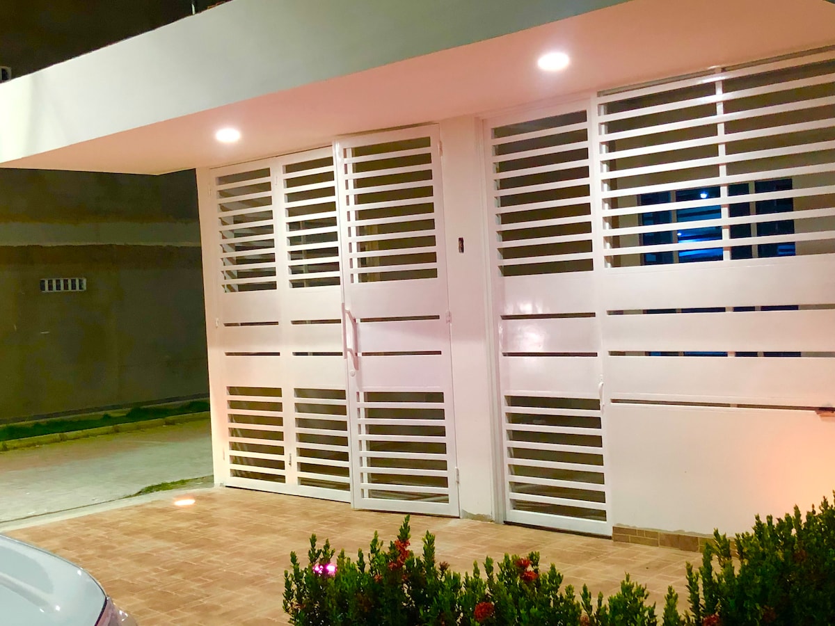 The entrance is framed by modern white slatted doors, providing a sense of openness. Soft lighting illuminates the outdoor area, accentuating the paved walkway and surrounding greenery. The clean lines of the architecture highlight the contemporary design of the home.