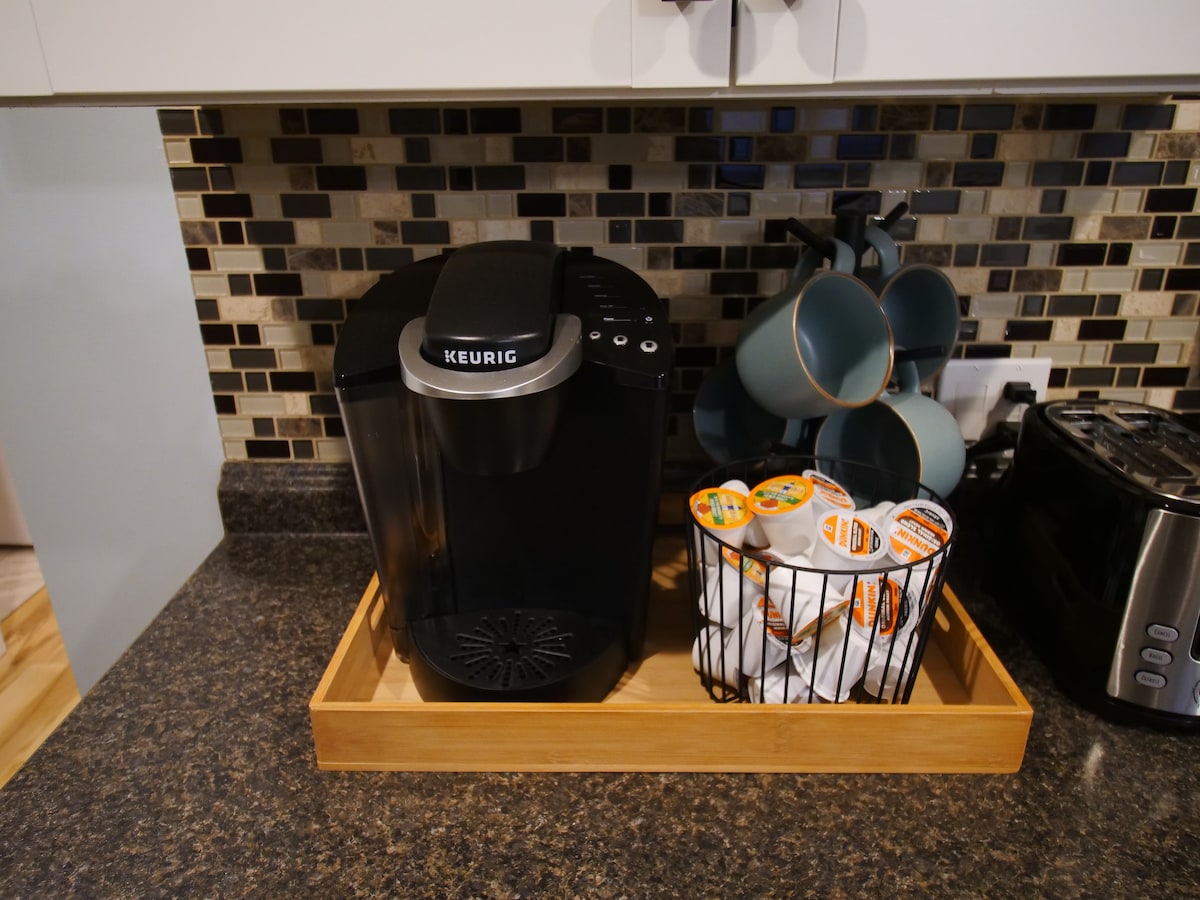 A modern kitchen countertop is shown featuring a Keurig coffee maker, surrounded by a collection of coffee pods organized in a wire basket. A wooden tray holds the coffee maker, while several teal mugs are neatly arranged in a utensil holder nearby.