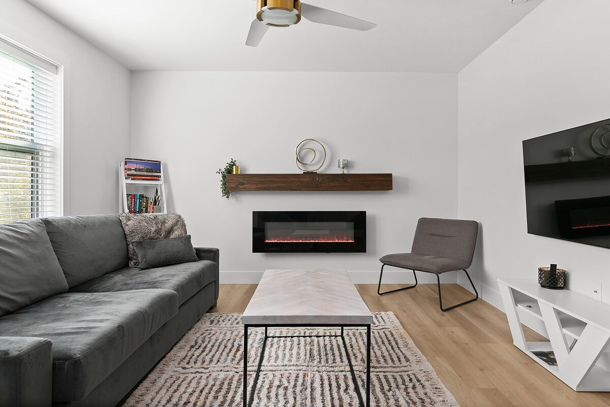 A modern living room features a grey sectional sofa with decorative pillows, complemented by a sleek coffee table. An electric fireplace is mounted on the wall beneath a floating shelf adorned with decor. A cozy armchair is positioned across from a wall-mounted television.