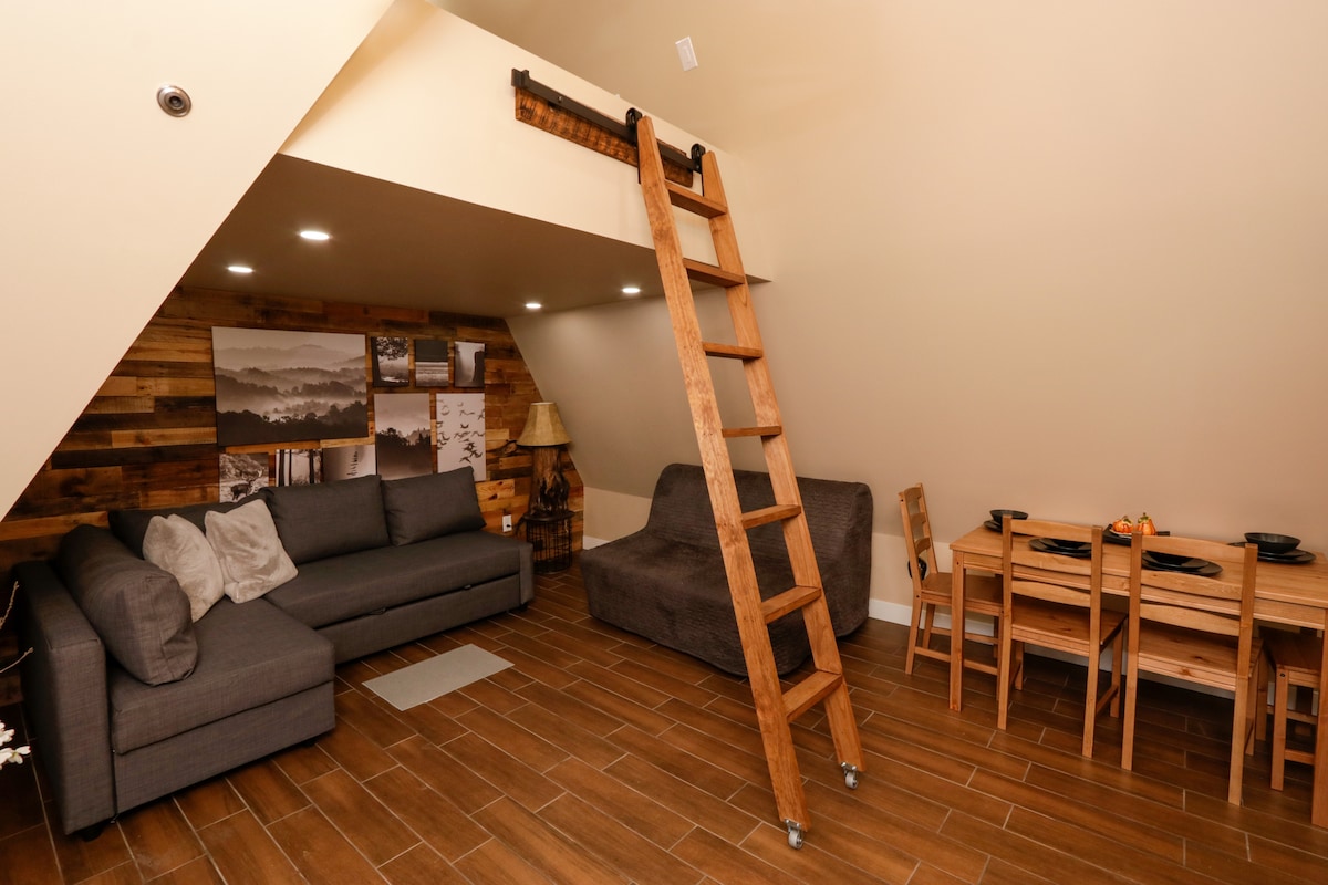 The interior space features a cozy seating area with two gray couches, one of which functions as a pull-out bed. A rustic wooden ladder leans against the wall, providing access to an elevated area. A small dining table is set for four, surrounded by wooden chairs.