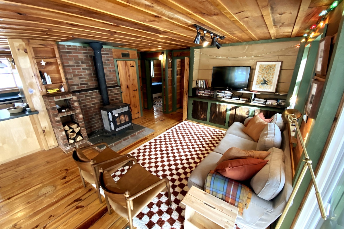 A comfortable living area is displayed with warm wooden walls and a cozy wood stove. A light-colored couch is positioned beside two stylish chairs. A patterned rug adds texture to the floor, while a small television and shelves are visible along the wall.