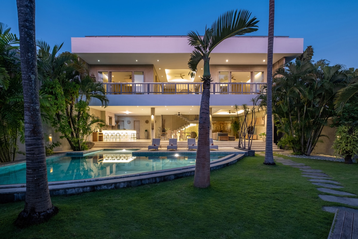 A spacious two-story villa is set within a lush garden, featuring palm trees and a well-maintained lawn. An inviting pool reflects the evening sky, while a large terrace overlooks the area. The open design offers views of the interior and outdoor spaces.