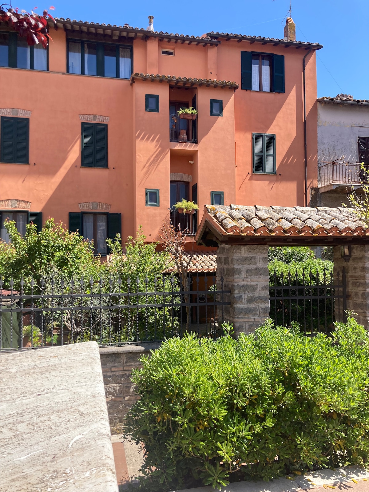 A colorful building with a terracotta facade is showcased, featuring multiple green-shuttered windows and flower boxes. A stone wall and wooden roof structure are visible in the foreground, surrounded by lush greenery that adds a touch of vibrancy.