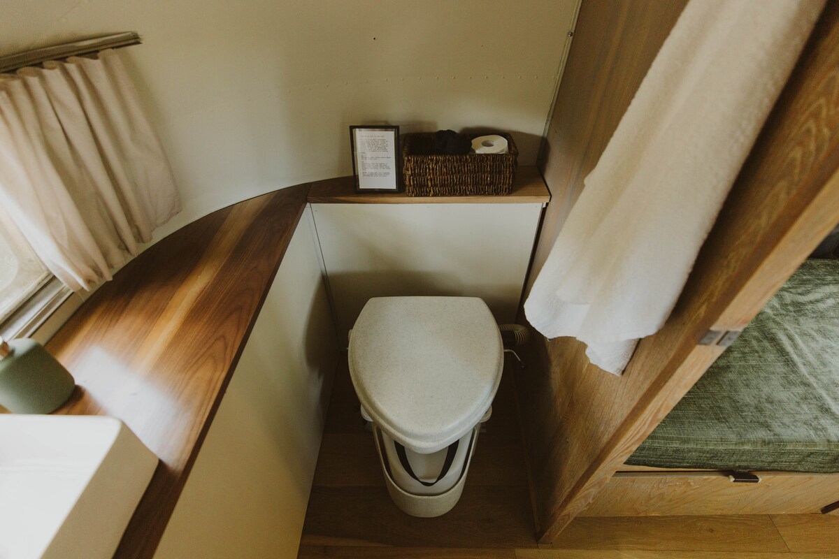The bathroom features a clean composting toilet positioned near a wooden countertop. A woven basket containing toiletries sits nearby, accompanied by soft white towels draped on the side. Natural light filters in through the window, enhancing the serene atmosphere.